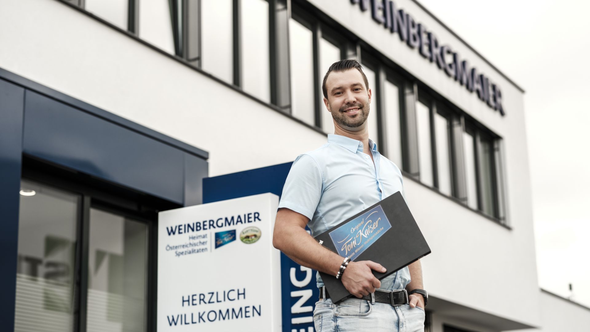 Ein junger Mann steht mit einer Toni Kaiser Mappe vor dem Weinbergmaier Firmengebäude