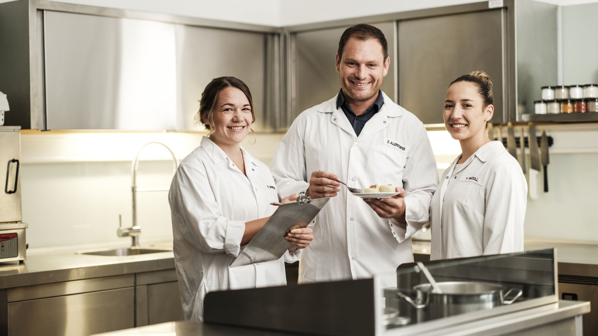 Ein Mann und zwei Frauen in weißen Kitteln stehen in der Küche